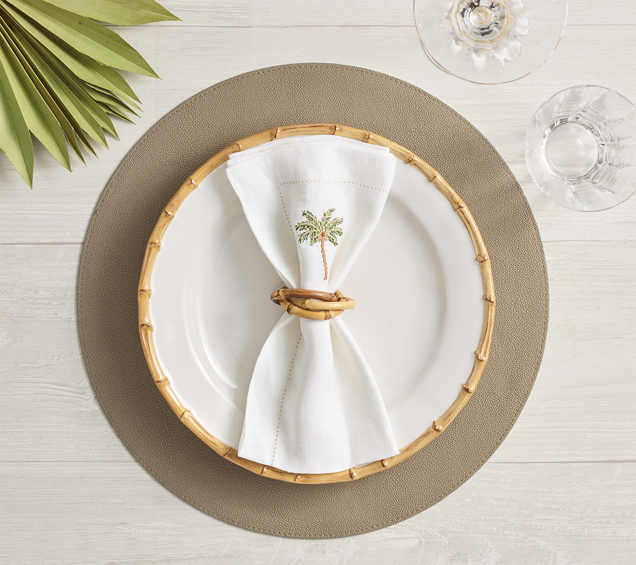 A white plate with a bamboo-style rim sits on a textured placemat. In the center, a white napkin is folded creatively and secured with the Kim Seybert Natural Bamboo Trinity Napkin Ring. Glassware accompanies the setting.