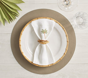 A white plate with a bamboo-style rim sits on a textured placemat. In the center, a white napkin is folded creatively and secured with the Kim Seybert Natural Bamboo Trinity Napkin Ring. Glassware accompanies the setting.