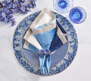A table setting with blue and white plates, a floral napkin ring, cobalt blue beads, a Kim Seybert Lagoon Placemat, a blue glass, and purple flowers arranged on a marble surface.