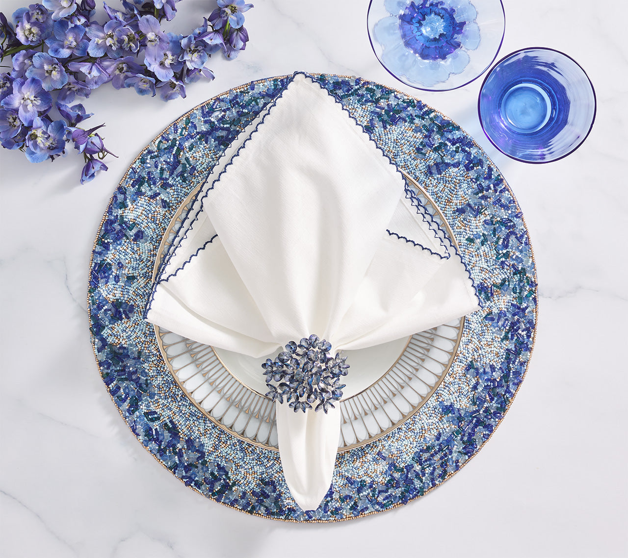 The Lagoon Placemat by Kim Seybert anchors a white napkin with a cobalt blue beaded ring on a patterned plate, alongside blue flowers and glassware—creating an ocean-inspired tablescape.
