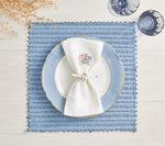 The Kim Seybert Vista Reversible Placemat in blue basketweave is styled with a white plate, blue-edged plate, knotted white napkin, two glasses, and dried stems on a white wooden table.