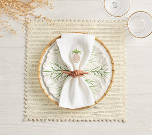 A decorative plate with a leaf pattern sits on a Kim Seybert Vista Reversible Placemat, topped with a white napkin featuring an embroidered pineapple and secured by a knotted brown napkin ring.