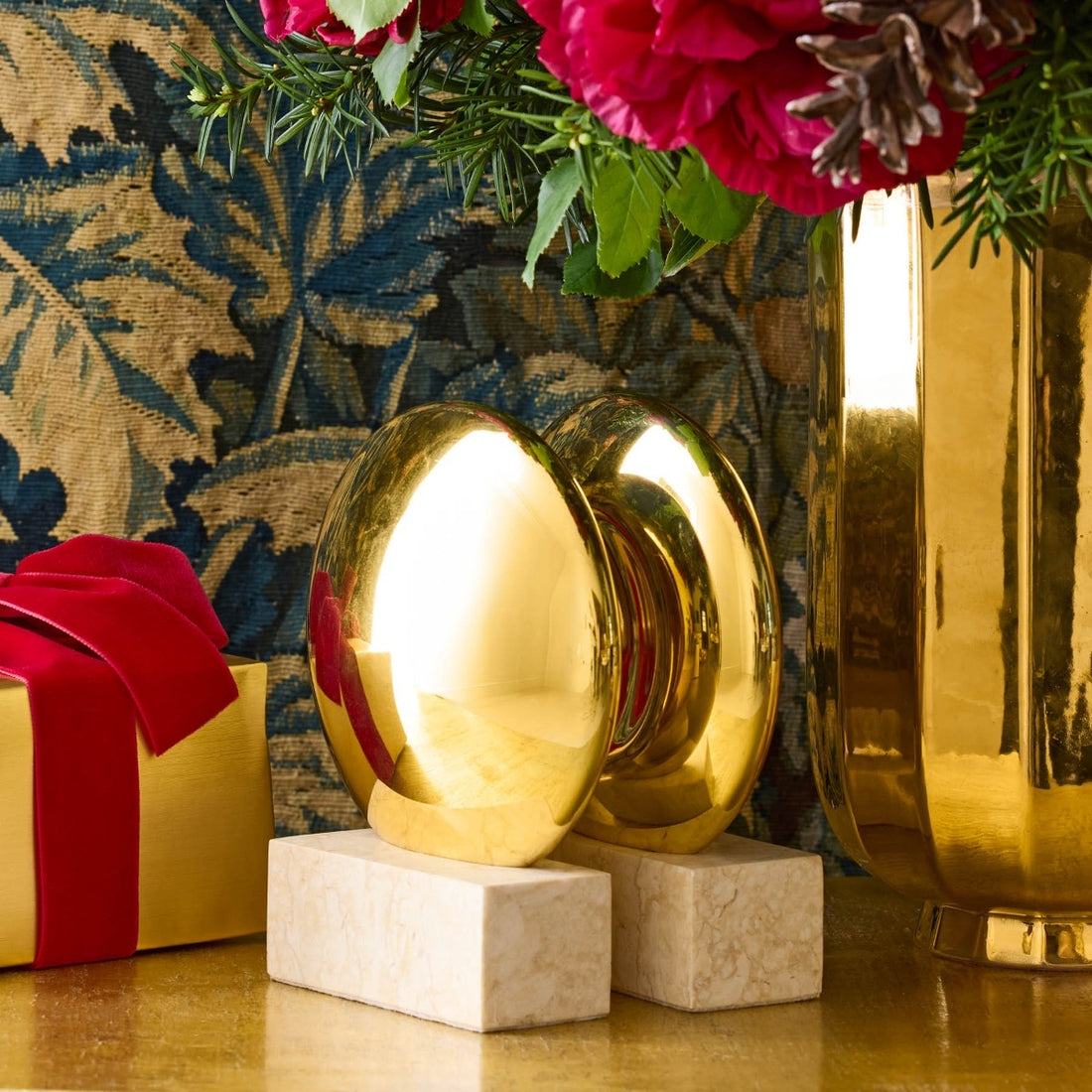 The Aerin Constantin Bookend Set, featuring shiny brass on marble bases, sits beside a gold vase with flowers and a gold gift box tied in red, arranged against a floral backdrop that reflects elegant Mid-century style.