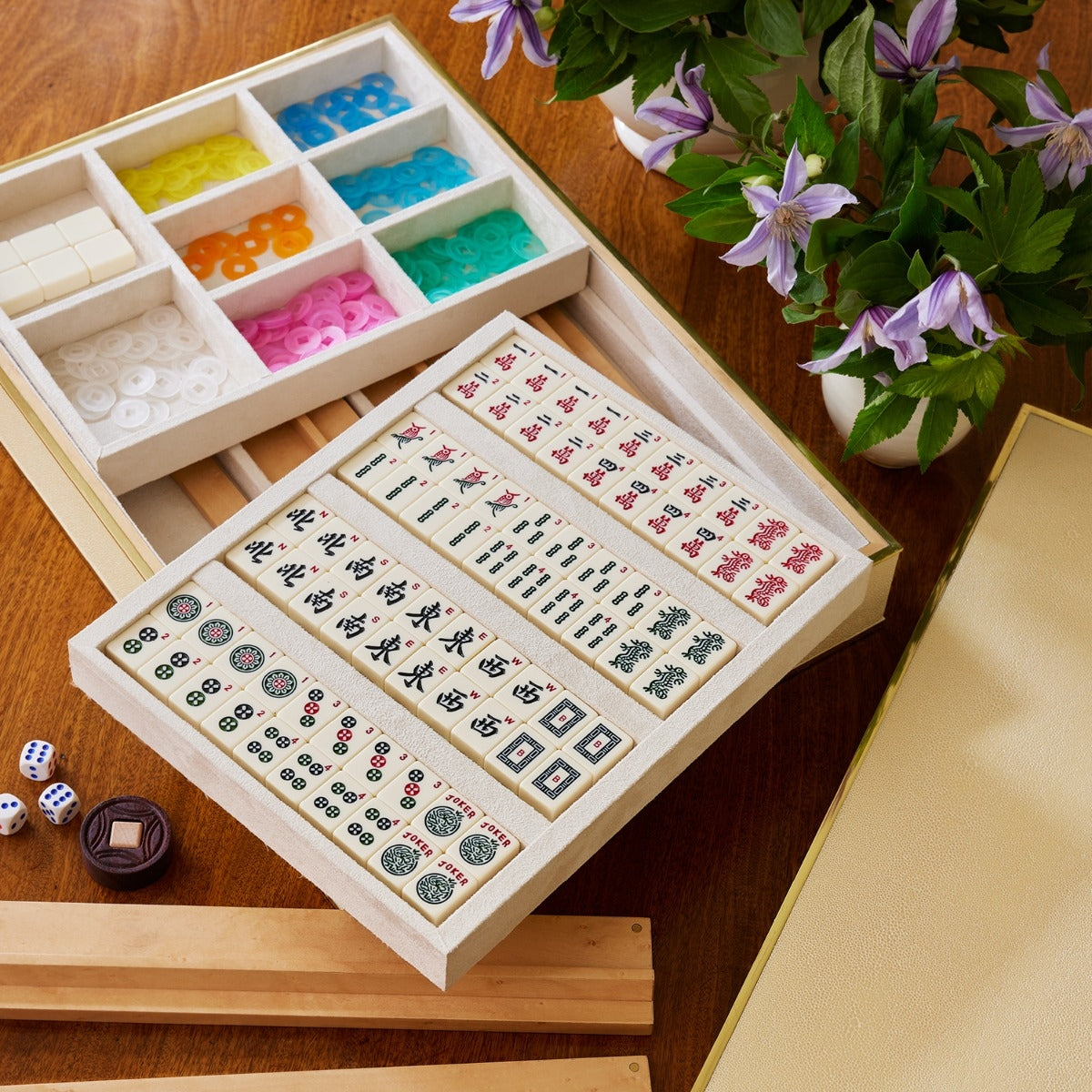 The Aerin Luxe Shagreen Mahjong Set in Cream, featuring engraved tiles, colorful chips, dice, and racks, is displayed on a wooden table beside a vase of purple flowers.