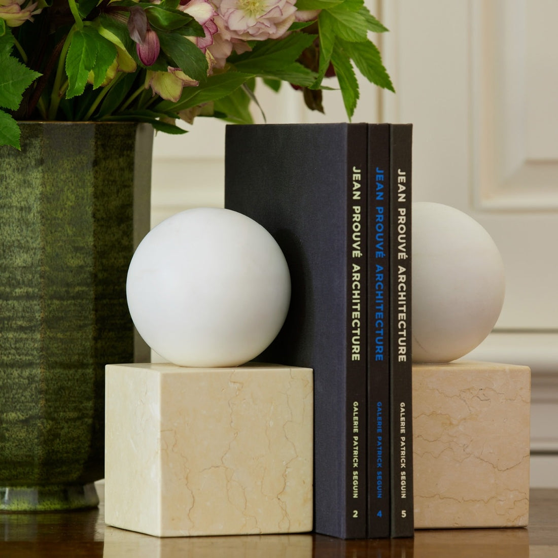 Aerin's Odette Bookend Set, featuring marble bookends topped with white spheres, holds three architecture books upright beside a green vase of leafy flowers, all arranged against a white background.