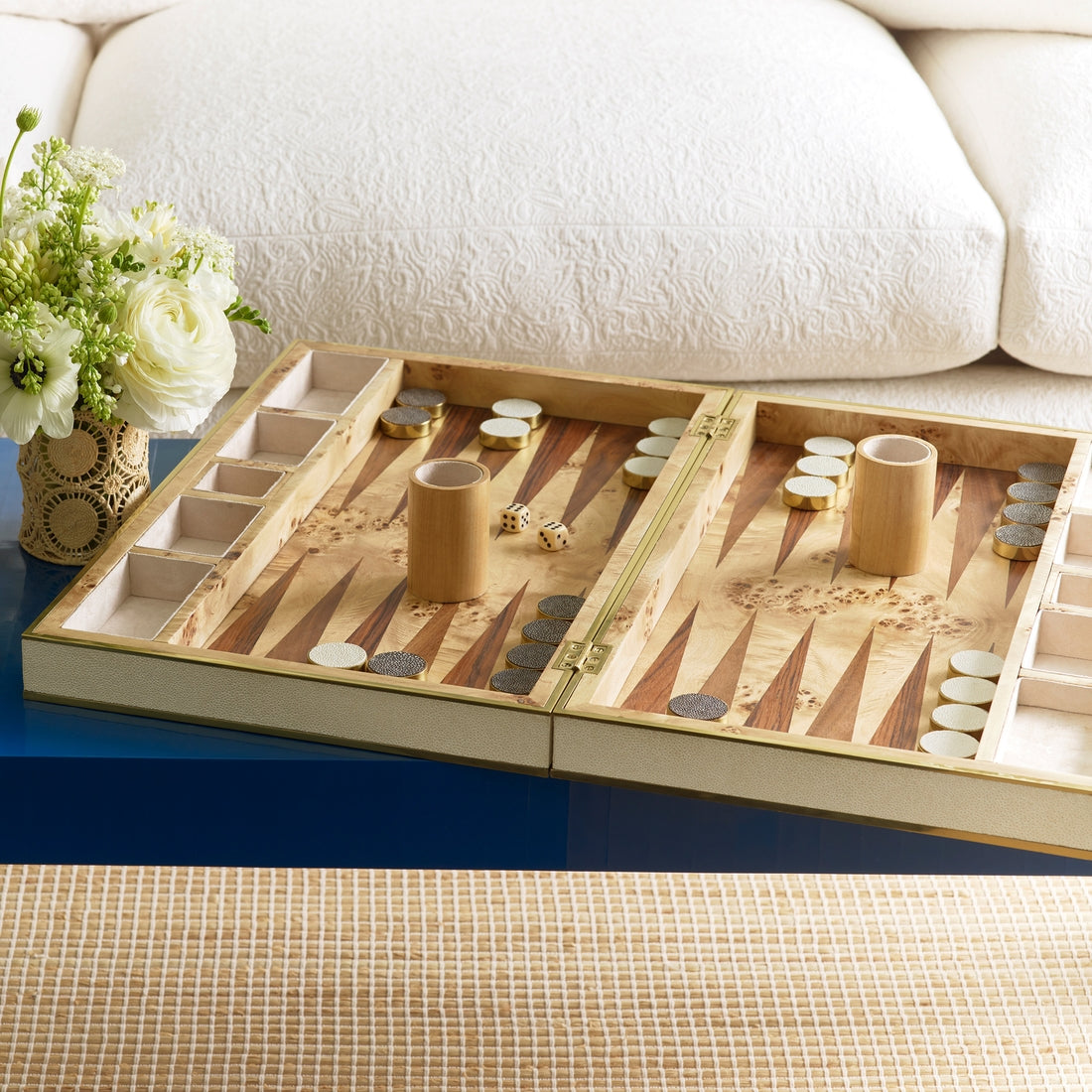 The Aerin Shagreen Backgammon Set in Cream, complete with playing pieces, dice, and cups, is displayed open on a blue table beside a vase of white flowers and a white couch in the background.