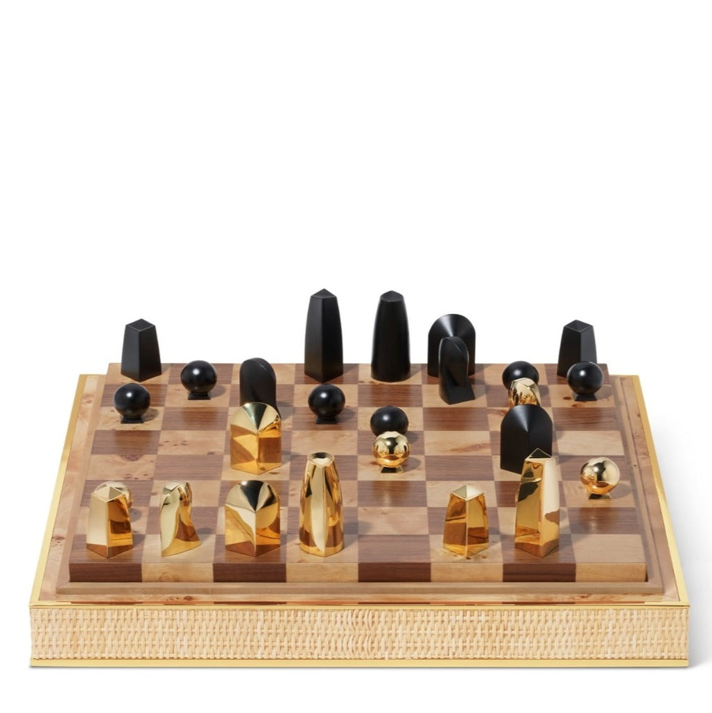 The Colette Cane Chess Set by Aerin features abstract gold-plated chess pieces in black and gold on a wooden board, mid-game, and comes with a stylish woven cane case.