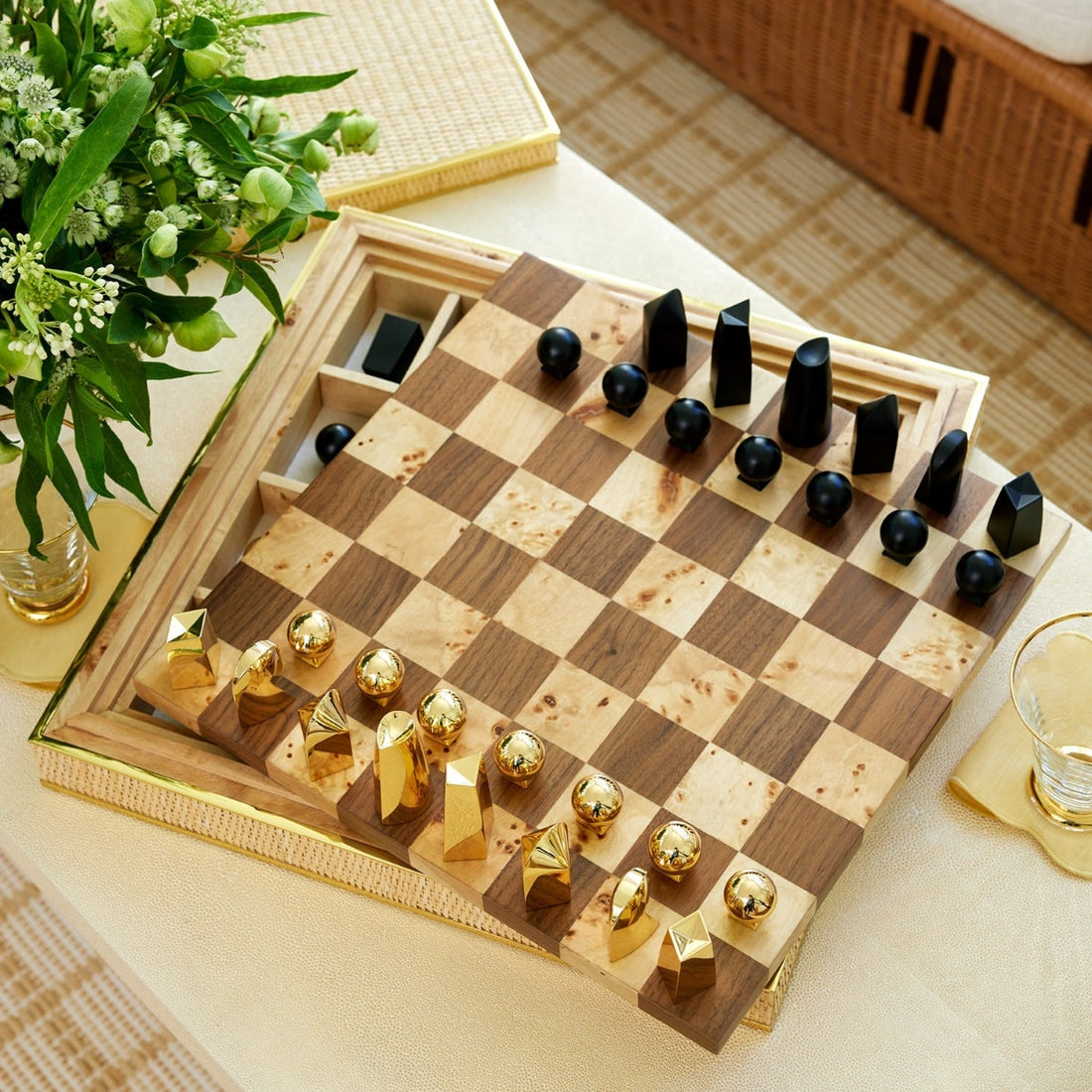 The Aerin Colette Cane Chess Set, featuring geometric gold-plated pieces and a wooden board, rests on a table with flowers, a glass of water, and its woven cane case for an elegant display.