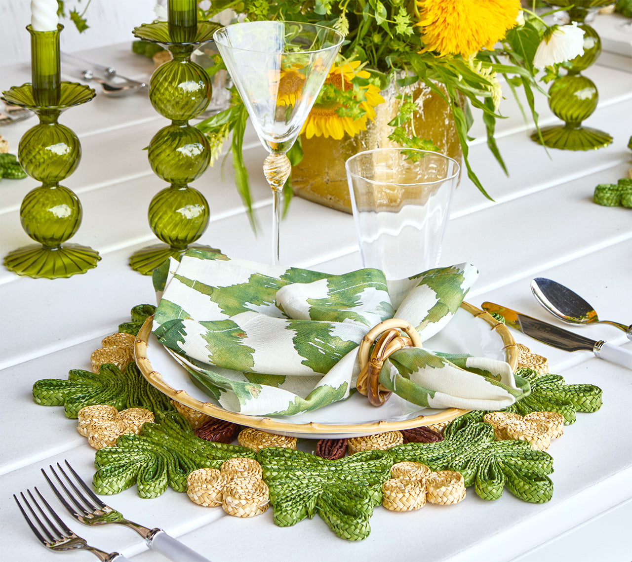 A table setting with green and gold accents features Kim Seybert's Verve Placemat in Green & Natural, a patterned napkin with a gold ring, elegant glassware, and a floral centerpiece with yellow flowers.