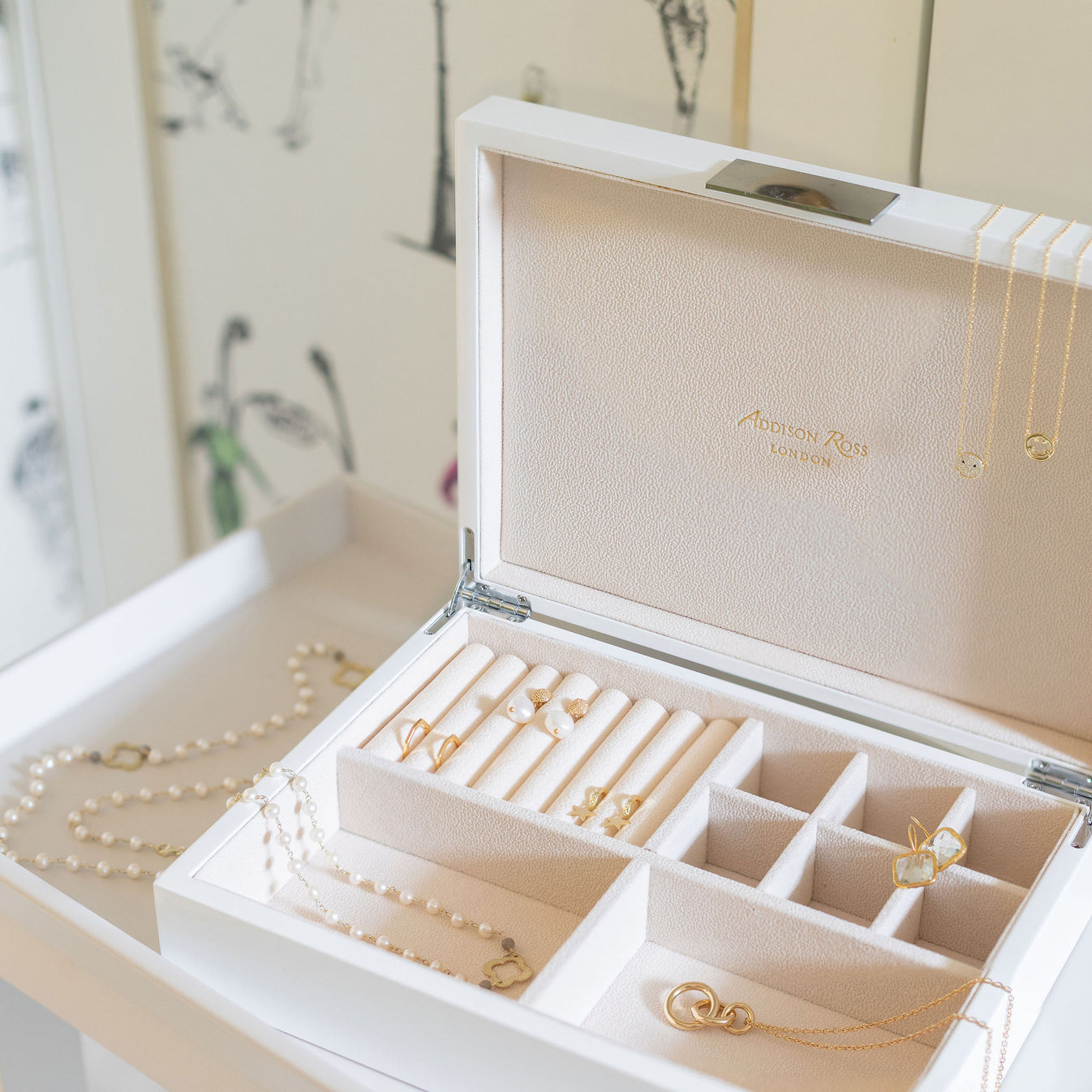 The Addison Ross Lacquer Jewelry Box with a polished white finish displays gold and pearl necklaces, rings, and earrings neatly organized in its compartments on a white dresser.