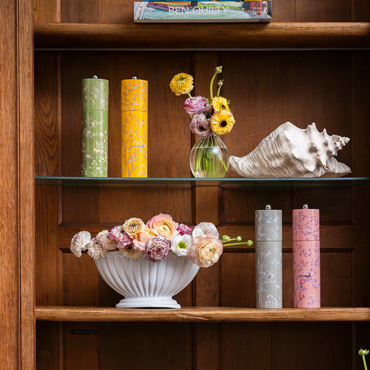 On this wooden shelf, vibrant vases and floral arrangements mix with two tall cylindrical containers featuring a ceramic grinder. An Addison Ross Chinoiserie Mill completes the display beside a large seashell and a cherished book by Sarah and David Ross.