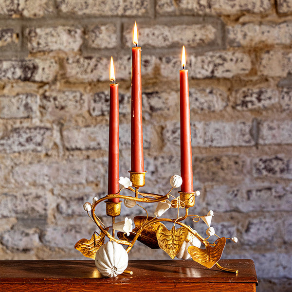 The Juliska Forest Walk Tabletop Collection features a gold candelabra with three red taper candles and oak leaf accents, displayed on wood against a textured brick wall.