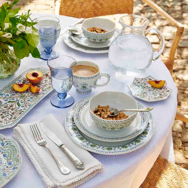 A round table set for two with Juliska’s Villa Seville Chambray Dinnerware Collection, bowls of granola, sliced peaches, blue glasses, coffee, a water pitcher, and a vase of flowers.