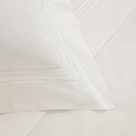 A close-up of a neatly made bed with a white pillow from Frette's Triplo Bourdon White/Milk Bedding Collection, featuring elegant three-line embroidery near the border, resting on matching cotton poplin white sheets.