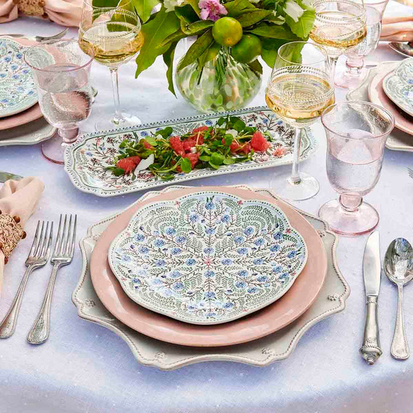 A table set with patterned plates from the Juliska Villa Seville Chambray Dinnerware Collection, pink and clear glassware, silver cutlery, a white tablecloth, a floral centerpiece, and a rectangular plate of salad.