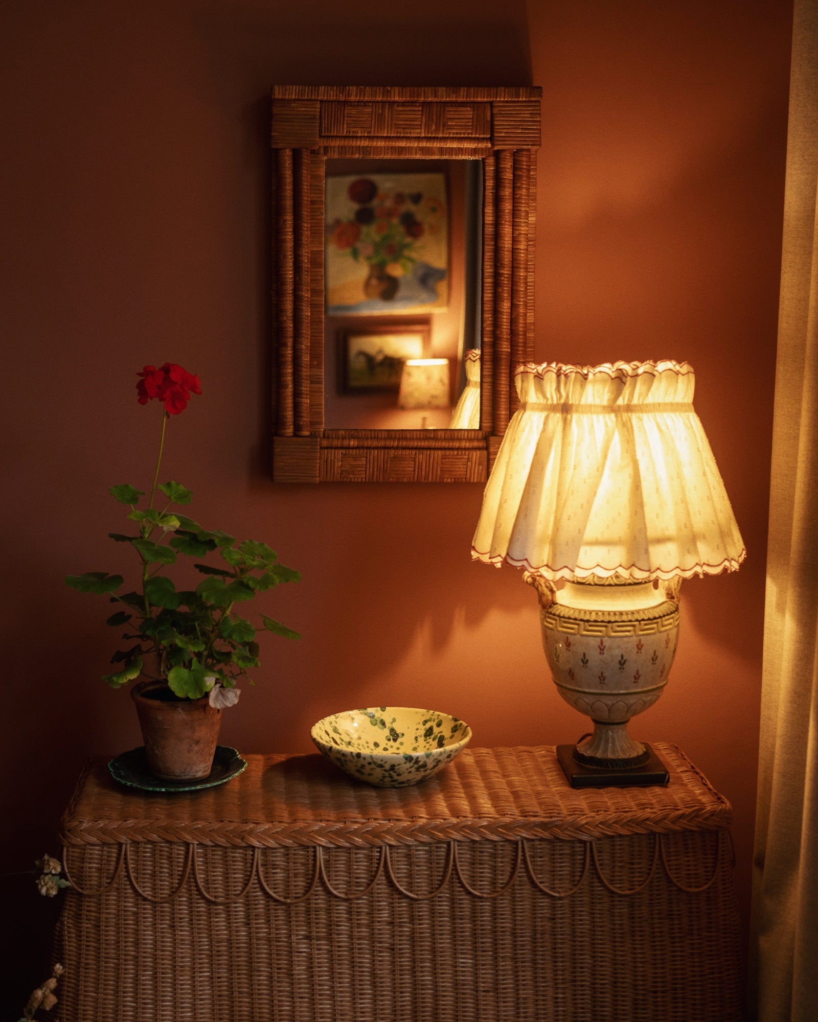 A wicker table with a potted red geranium, decorative bowl, and vintage lamp sits against a brown wall with a framed Sharland England Column Mirror reflecting wall art.