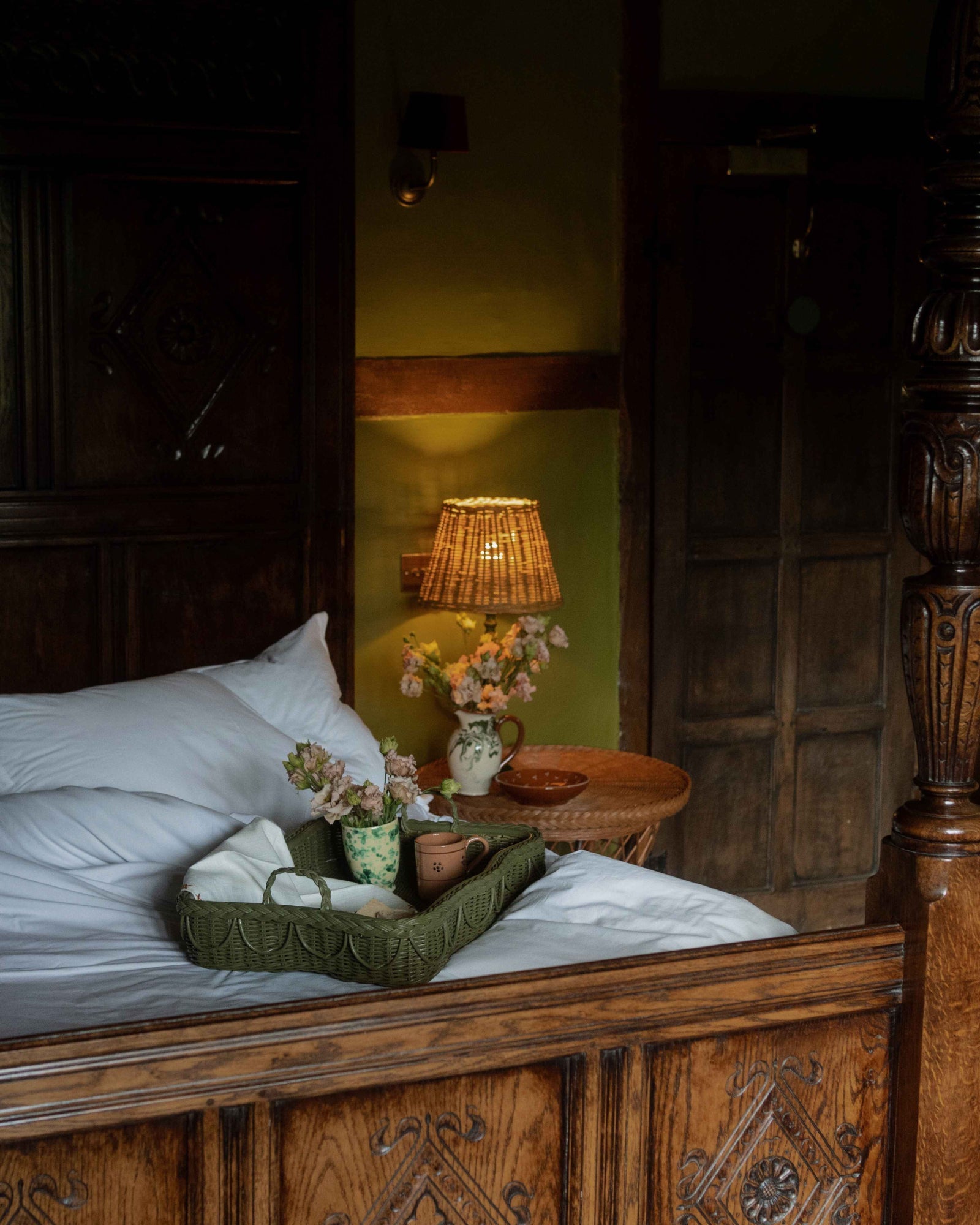 A cozy bedroom features an ornate wooden bed, white pillows, a Sharland England Hadley Tray Collection tray with tea and flowers, a wicker side table, and a lit lamp against a green wall.