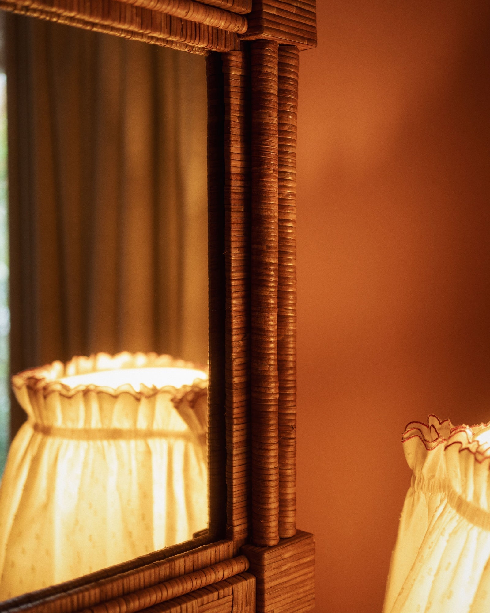 A ruffled lampshade is reflected in the Sharland England Column Mirror, featuring a wooden frame, set against a warm brown wall with curtains in the background.
