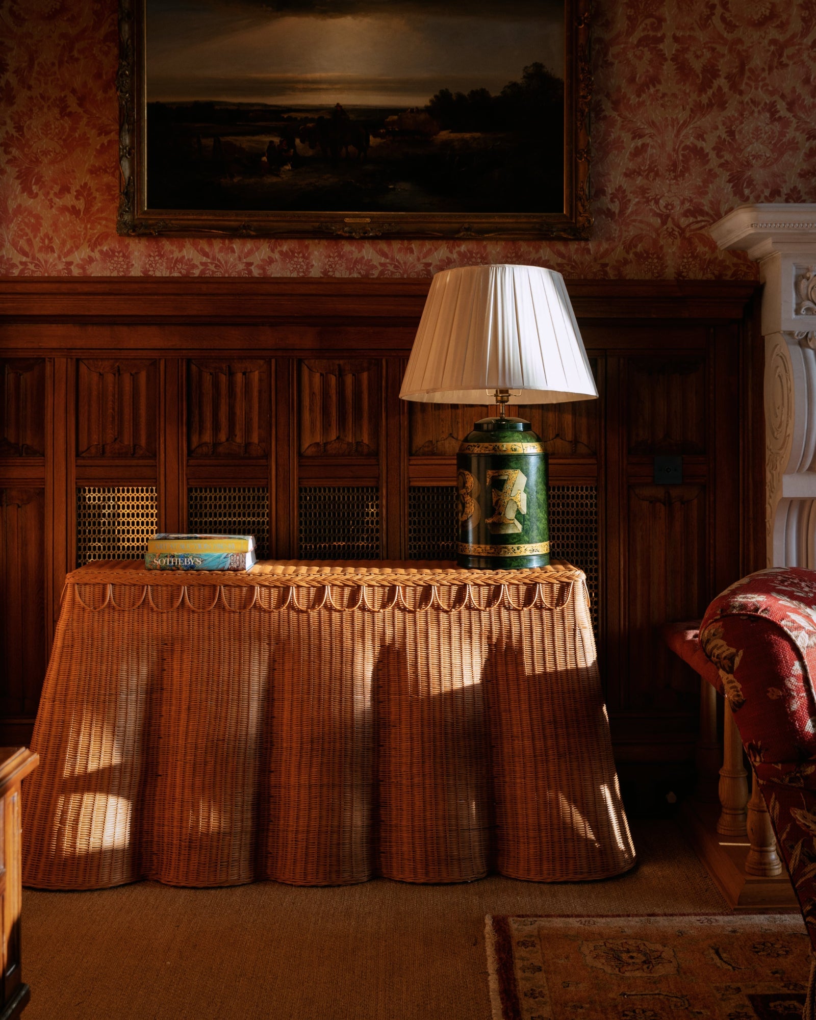 The Adeline Console Table by Sharland England, styled with books and a lamp, is placed against a wood-paneled wall beneath a framed painting in a traditionally decorated room.
