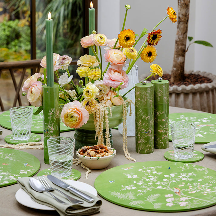 The table setting includes green floral placemats, green candles, various flowers in a vase, glasses, and a bowl of nuts. The Addison Ross Chinoiserie Salt and Pepper Mill Collection subtly enhances the ambiance with its exquisite charm.