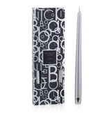 A silver tapered candle is displayed next to its box, adorned with a black and white letter pattern design, ready to elegantly complement any candlestick from the Baobab Collection Candela by Baobab.