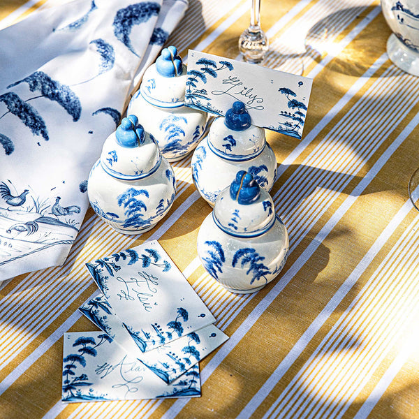 Juliska's Country Estate Place Card Holder Set, featuring Delft Blue miniature jars, is displayed on a yellow striped tablecloth with handwritten names. Blue-patterned linen adds a charming touch to the scene.