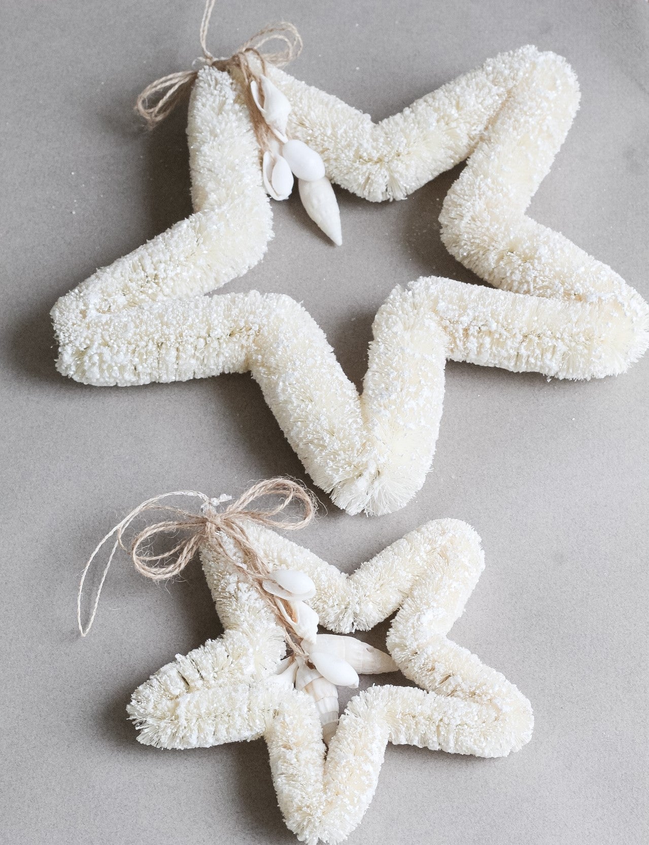 The Dekorasyon Bottlebrush and Shell Star Ornament features two white star-shaped designs crafted from textured abaca fiber, adorned with twine bows and decorative white shells for a charming coastal touch.