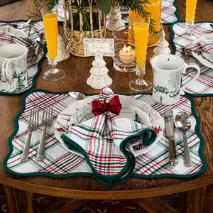 A festive table features plaid placemats, Berry & Thread holiday plates and mugs, silverware, napkins tied with red bows, orange drinks, and a “Sarah” place card in Juliska’s Berry & Thread White Tree Place Card Holder Set.