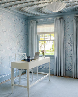 A home office with light blue floral wallpaper, a white desk with a laptop, a white chair, blue drapes, and a large window letting in natural light, accented by the Conrad Desk from Made Goods.