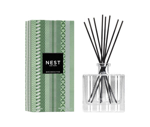 The Nest Birchwood Pine Collection reed diffuser features a patterned green box and clear glass bottle with black reeds, capturing the scent of a winter forest.