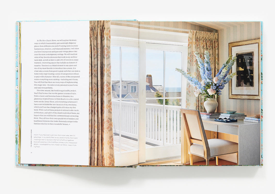 An open book, "The New Classic Home" by Abrams, displays a page of text on the left and a photograph on the right, featuring a sunlit office with classic and modern design elements that overlooks a balcony.