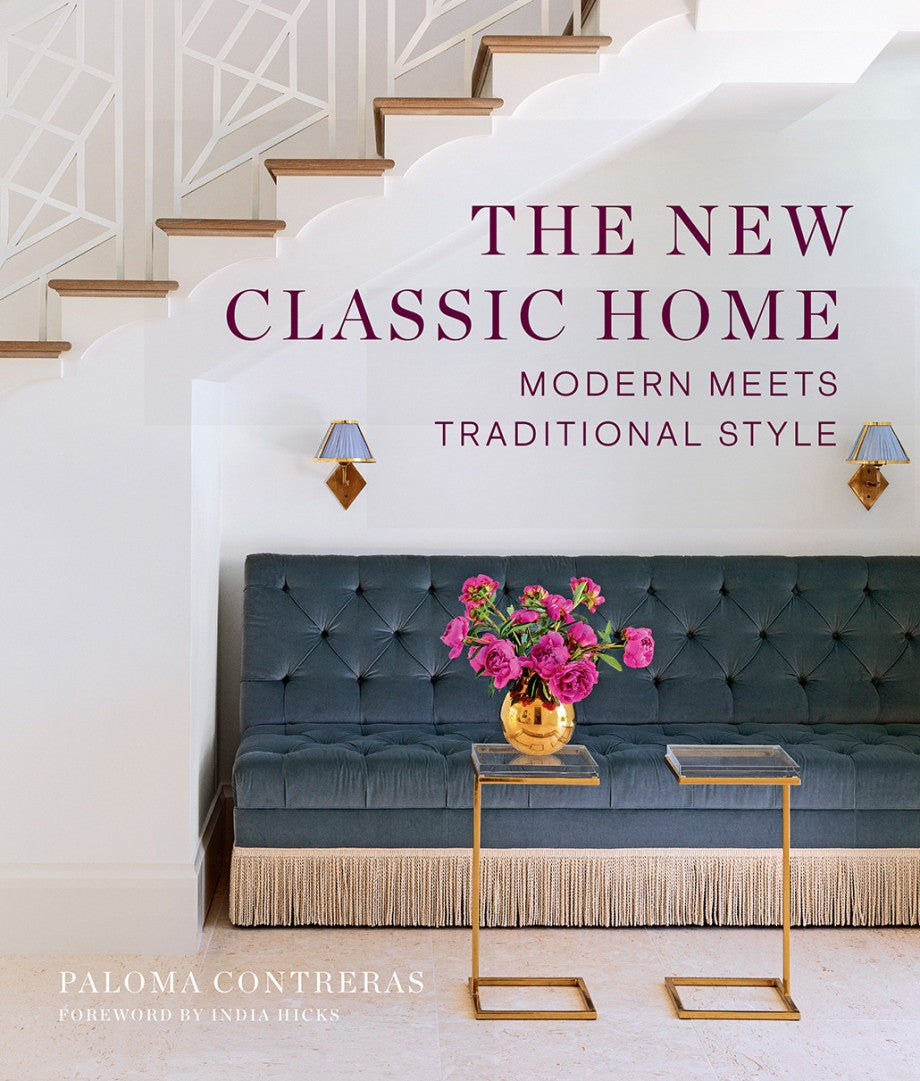 Cover of "The New Classic Home" by Abrams features classic home design with a tufted bench with fringe, gold tables, pink flowers, wall sconces, and a staircase with a modern railing.