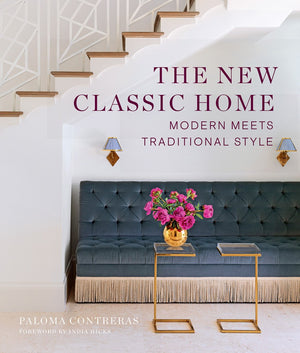 Cover of "The New Classic Home" by Abrams features classic home design with a tufted bench with fringe, gold tables, pink flowers, wall sconces, and a staircase with a modern railing.