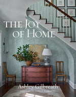 Book cover of "The Joy Of Home" by Common Ground, featuring an entryway with a red dresser, two chairs, a lamp, and a large potted plant. The elegant style is complemented by a staircase and framed artwork in the background, epitomizing Southern decorating charm.