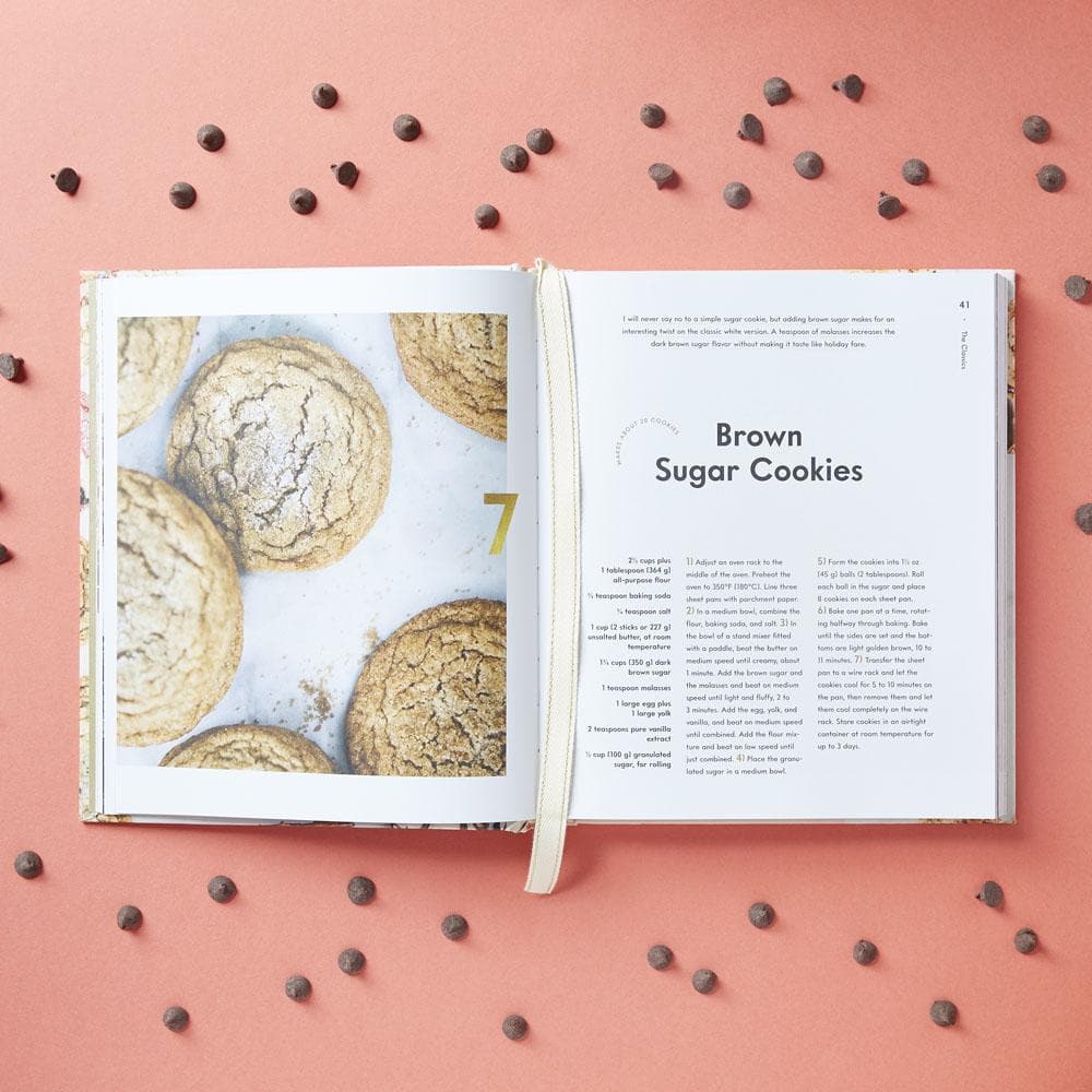 An open 100 Cookies book by Chronicle Books displays a brown sugar cookies recipe and a full-page photo, with chocolate chips scattered on pink—a tempting preview of the delicious cookie recipes inside.