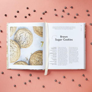 An open 100 Cookies book by Chronicle Books displays a brown sugar cookies recipe and a full-page photo, with chocolate chips scattered on pink—a tempting preview of the delicious cookie recipes inside.
