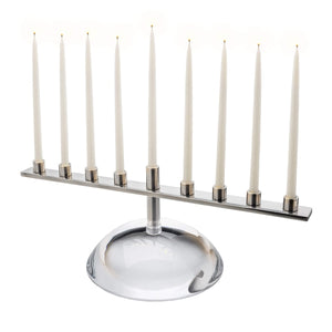 The Riverstone Menorah by Simon Pearce features a modern, handmade design with nine lit white taper candles on a reflective glass base, adding nature-inspired elegance to any space.
