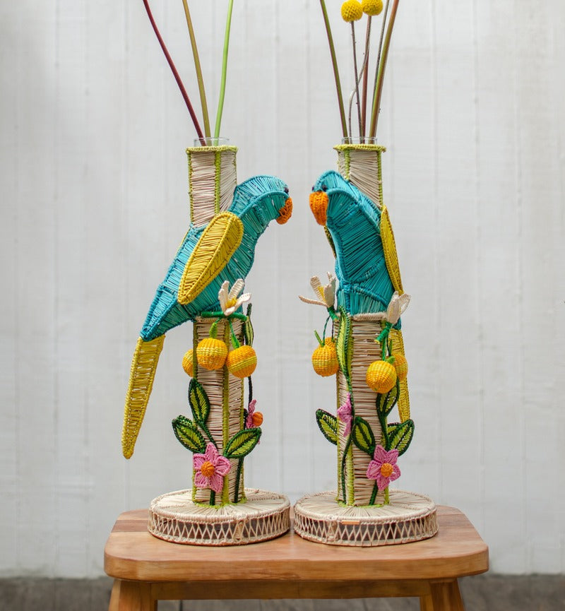 The El Pajaro Flower Vase by Mercedes Salazar features colorful woven parrots atop tall bases with floral and leaf details, shown on a wooden stool against a white background for a vibrant tropical accent.