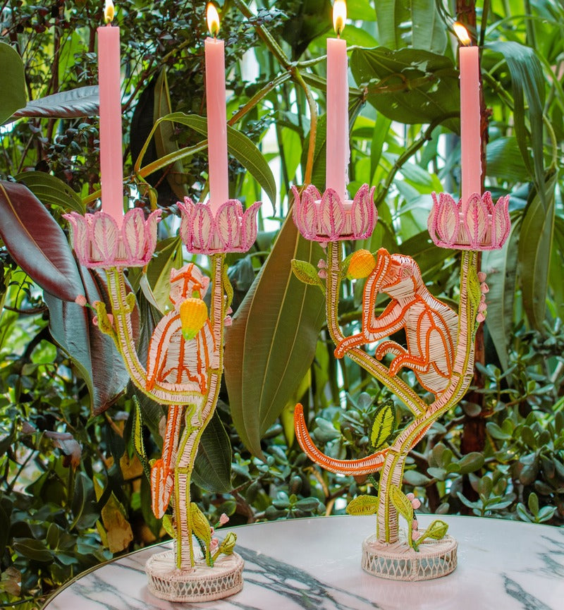 Two whimsical El Mico Candle Holders by Mercedes Salazar, shaped like monkeys and each holding three pink candles, rest on a marble table surrounded by lush green plants.
