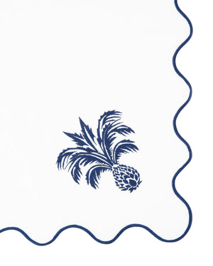 The Ananas Blue Napkin Set by Aquazzura Casa features white Italian linen napkins with a navy blue embroidered pineapple and palm tree in one corner and a scalloped edge trimmed in navy blue.