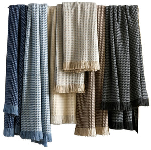 The Cleo Throw Collection by Matouk features five textured, woven blankets in blue, light blue, cream, brown, and black with fringed ends and a classic basketweave pattern for an elegant look.