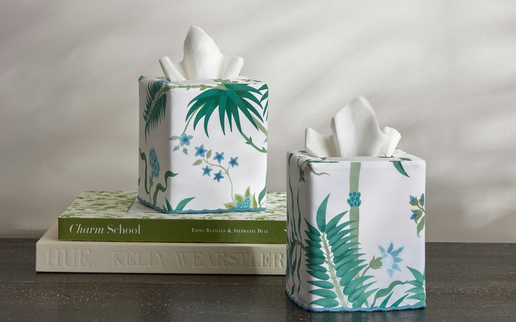 Two Matouk Palmyra Tissue Box Covers in Emerald, featuring botanical prints, bring island-inspired decor to a dark table; one sits atop a stack of three books.