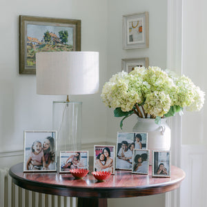 A round wood table with Addison Ross’s Cane Silver Frame Collection displays family photos, a lamp, two red bowls, and a white hydrangea vase, set against a white wall decorated with framed artwork.