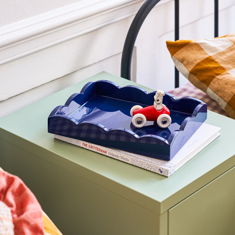 A small red toy car sits on "The Gritterman" by Orlando Weeks, arranged on an Addison Ross Scalloped Navy Lacquer Tray from Addison Ross. These items rest on a green nightstand beside a bed with checkered pillows, enhancing the cozy bedroom's charm and character.