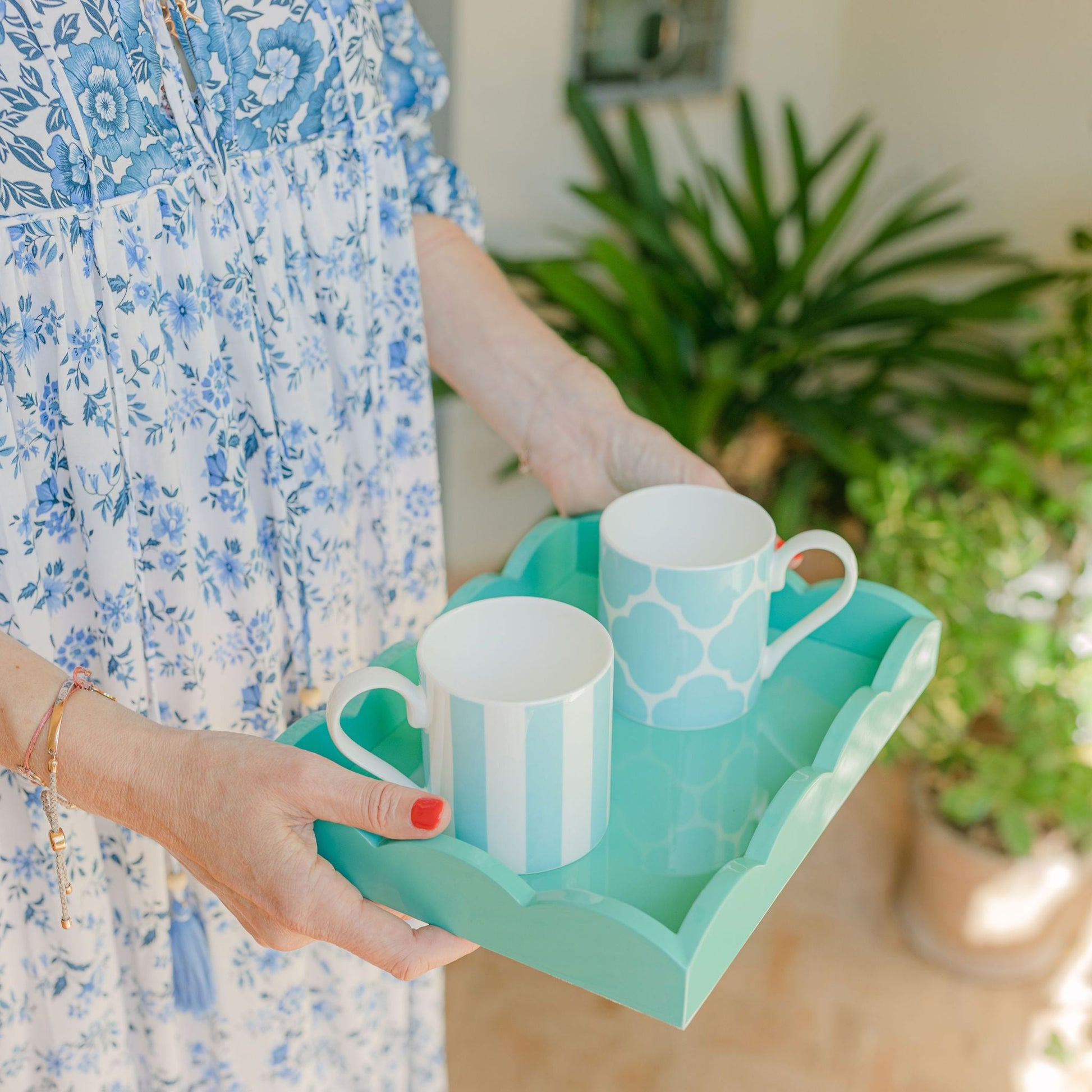 A person holds an Addison Ross Scalloped Turquoise Lacquer Tray from the Addison Ross collection, showcasing two patterned mugs—one striped and the other with a geometric design. Dressed in a blue floral dress, they are surrounded by lush greenery.