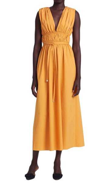 A woman stands exuding modern femininity in a sleeveless, yellow Altuzarra Fiona dress from Italy, featuring a mid-length cut with a ruched waistband and v-neckline, paired with black shoes.