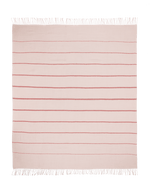 A square, light pink Matouk Amelia Stripe Throw with evenly spaced horizontal red stripes and white fringe on two edges is laid flat on a green background.