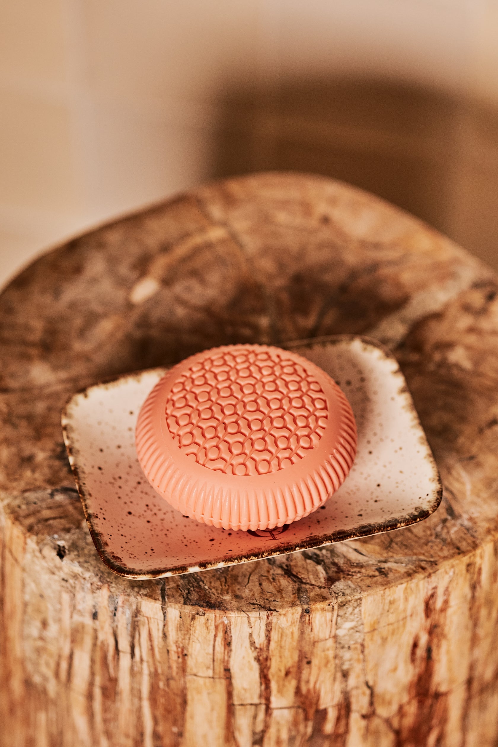 The Baobab Collection Women Body Wellness Collection coral-colored, textured soap with a delicate floral scent sits on a white ceramic dish atop a wooden surface.