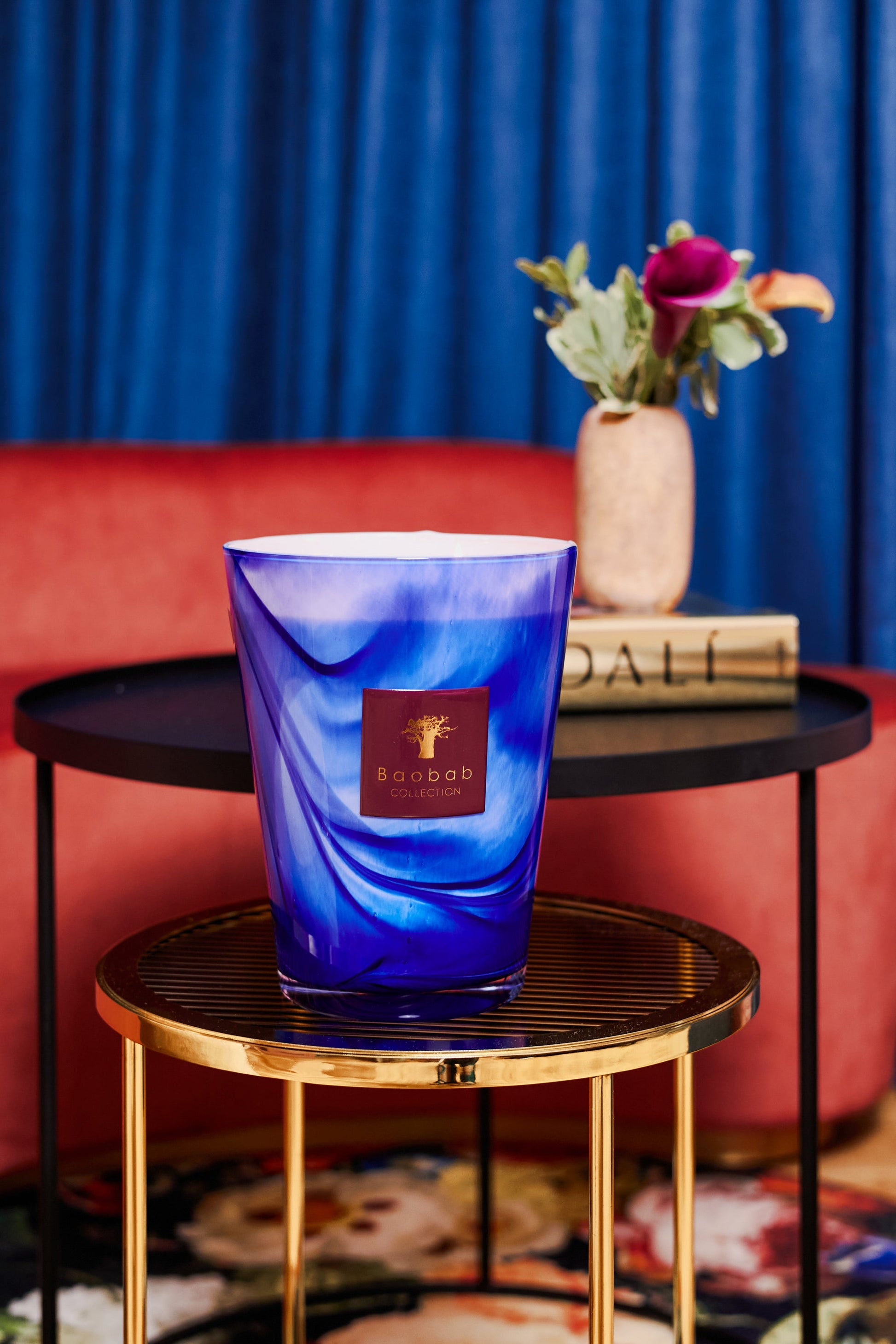 The luxurious ambiance is enhanced by a Baobab Collection Atlas Majorelle Candle resting on a gold stand in front of a plush red couch and black round table. The air is filled with the rich scent of patchouli, while blue curtains softly drape in the background.
