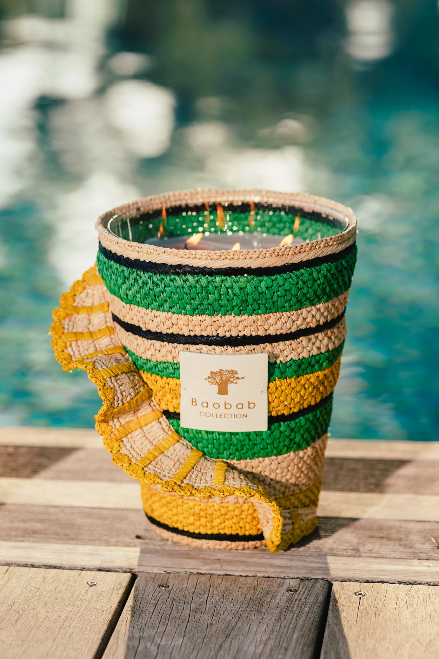 A woven candle holder with green, yellow, and natural stripes from Baobab's "Fety Milalao Candle" sits on a wooden surface near a pool. Flames flicker inside, releasing vetiver and ylang-ylang scents.